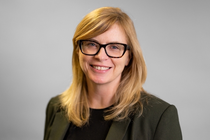 Close up photo of Anne Beath with a grey jacket and long blonde hair and glasses on a grey background